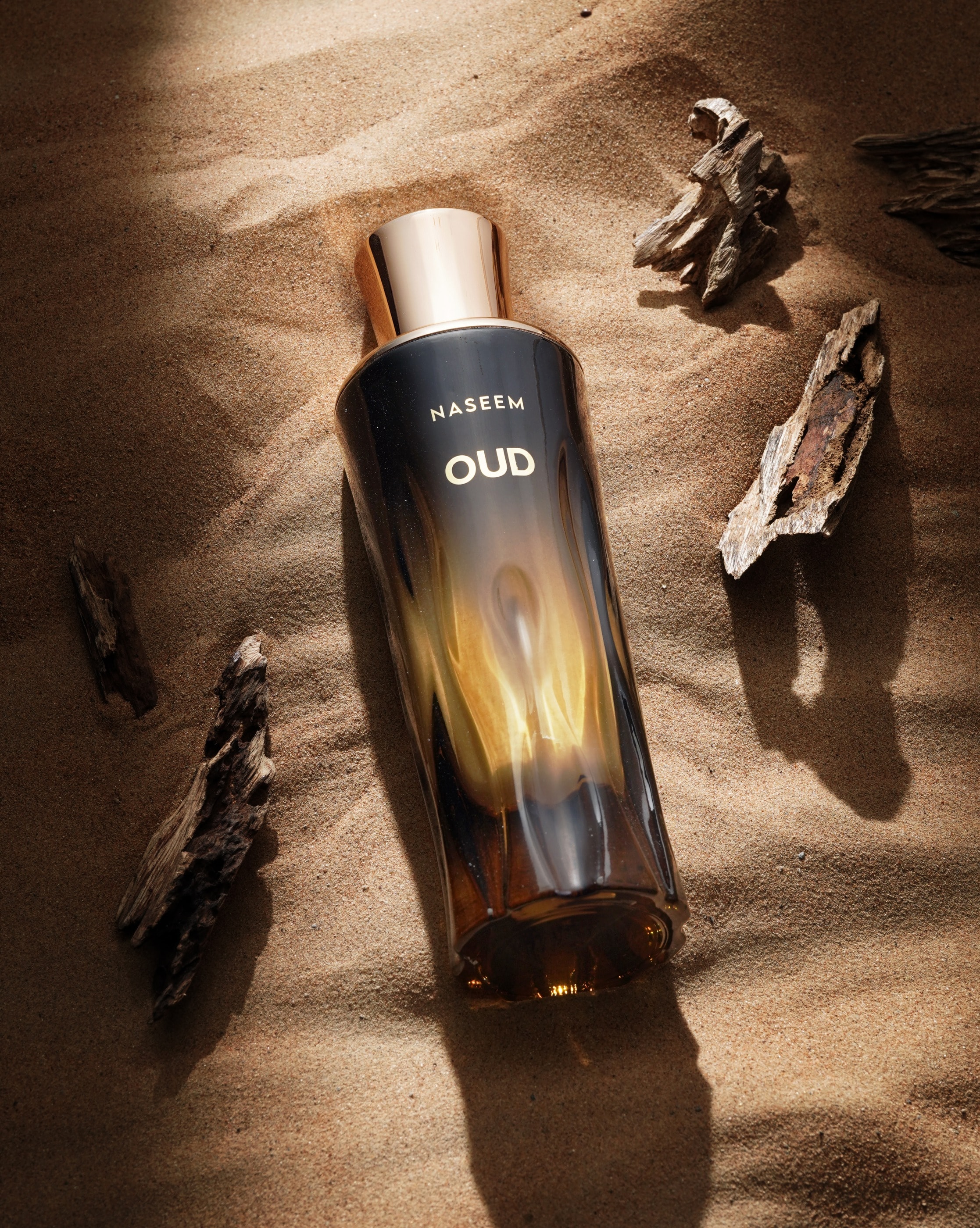 Een flacon Oud van Naseem op golvend woestijnzand, tussen stukjes agarhout, in warm laag zonlicht